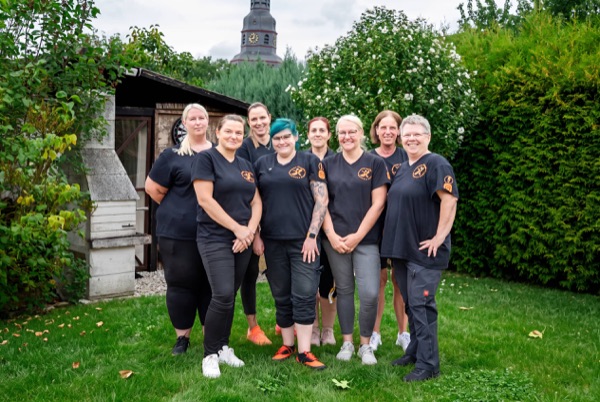Acht Kolleginnen in dunklen Dienstshirts mit AP-Ruth-Logo stehen im Garten, hinter ihnen ein Kirchturm