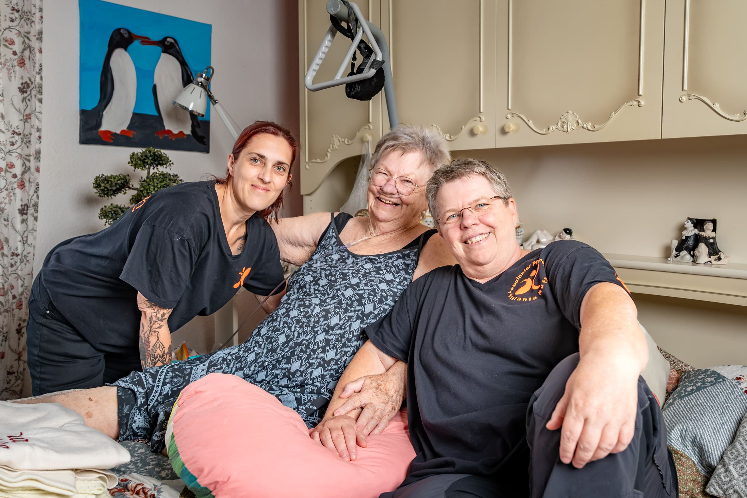 Zwei Pflegekräfte in dunklen Dienstshirts mit AP-Ruth-Logo sitzen lächelnd mit einer Kundin auf dem Bett im Schlafzimmer