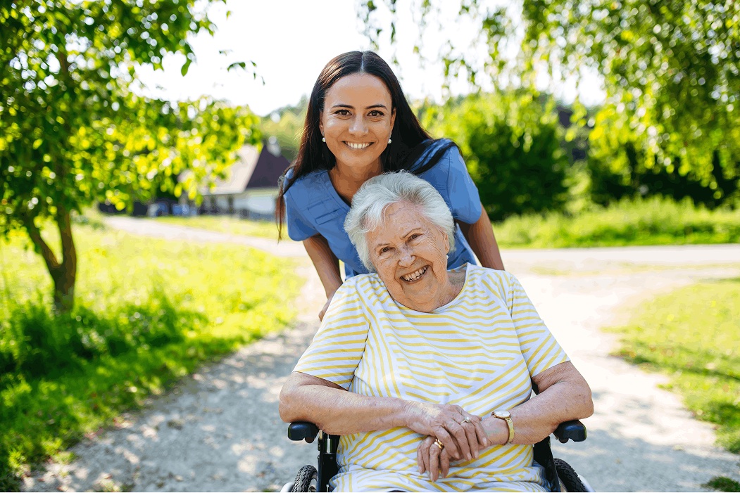 Eine Pflegerin schiebt eine lachende Seniorin im Rollstuhl über einen Weg